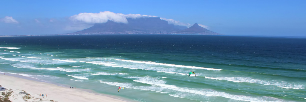 Turquoise waves along a Cape Town beach with Table Mountain and Lion’s Head in the distance under a clear blue sky. Showing the natural beauty that inspires the Den of Luxe founder.